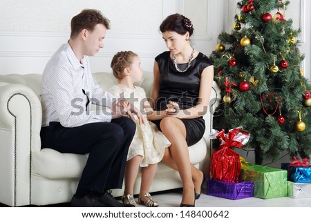 Father and mother talk with daughter on sofa near Christmas tree in light room.