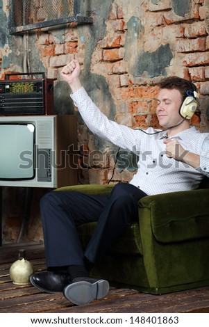 Happy dancing man in big headphones listens old radio and sits in armchair in very old house.