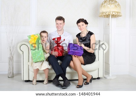 Father, mother and daughter sit at white sofa with bright gifts in light room.
