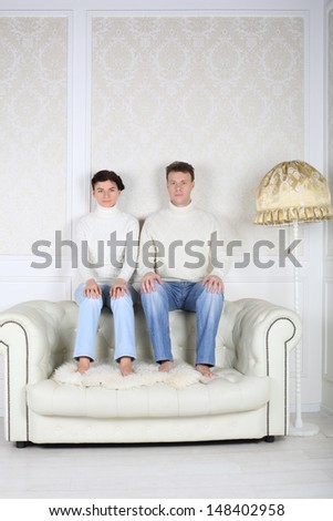 Serious husband and wife sits on back of white leather sofa at home.