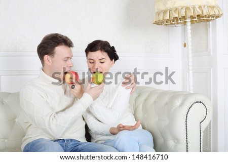 Happy husband and wife bite fruit and sit on white sofa in white room at home.
