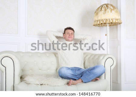 Relaxed barefoot man in white sweater sits on white sofa at home.