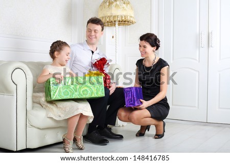 Father and mother give gifts to little daughter at sofa in light room.