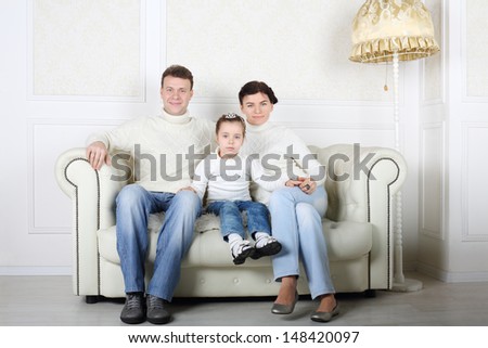 Happy family in white sweaters and jeans sit on white sofa in white room at home.