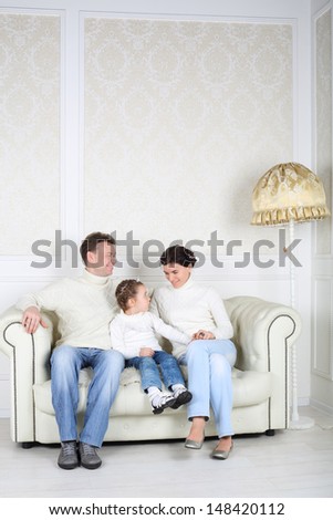 Family in white sweaters and jeans sit on white sofa in white room at home. Father and daughter look at mother.