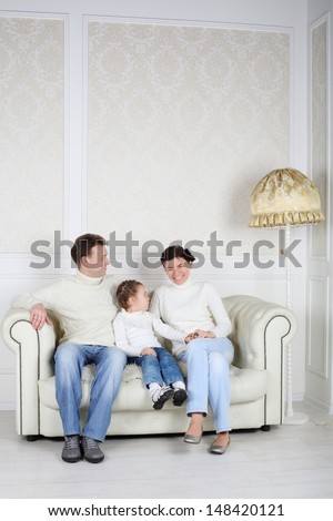 Family in white sweaters and jeans sit on sofa at home. Father and daughter look at smiling mother.