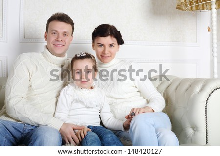 Father, daughter and mother in white sweaters and jeans sit on sofa at home.