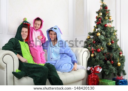 Family in colorful costumes of dragons sit on sofa near Christmas tree.