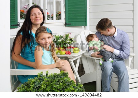 Happy mother with daughter and father with son sit at white table next to their house. Focus on woman and girl.