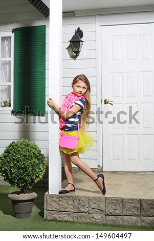 Little girl with pink handbag stands on white porch of house.