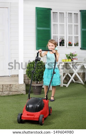 Happy cute little girl mows lawn by red lawn mower next to white house.