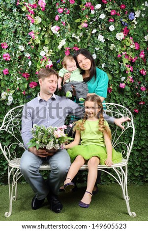 Happy family of four sit on white bench with bunch of flowers and stand behind in garden near verdant hedge.