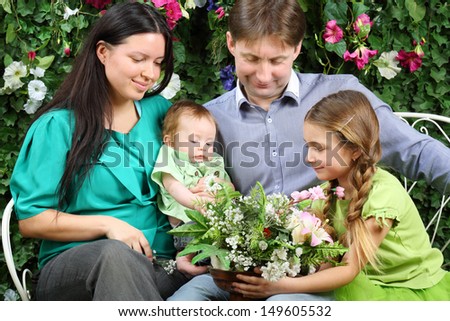 Father, mother, sister and baby look at bunch of flowers on bench in garden near verdant hedge.