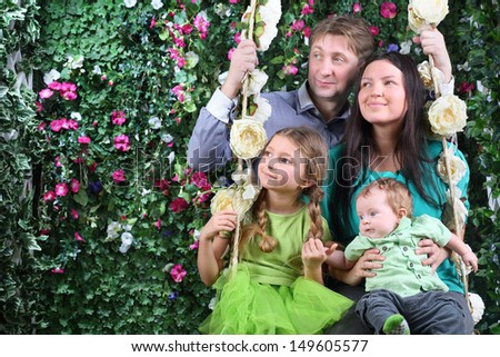 Happy family of four on swing look at distance near hedge with flowers in garden.