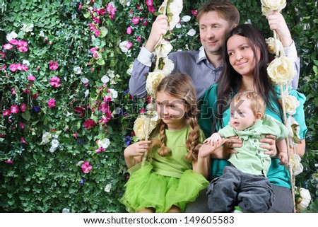 Happy family of four on swing look toward near hedge with flowers in garden.