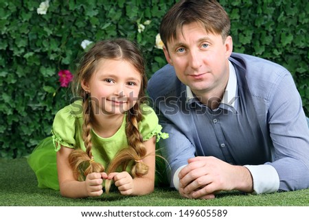 Father and little daughter lie on green grass near hedge in garden.