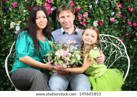 Happy family of three sit on bench, hold flowers and look at camera in garden near verdant hedge.