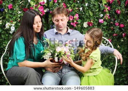 Happy family of three sit on white bench and hold flowers in garden near verdant hedge.