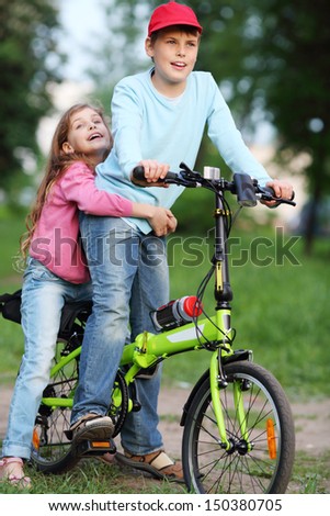 Happy brother and sister ride bicycle and sing in park at summer day.