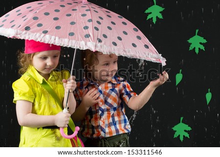 Little girl and boy hiding under a pink umbrella against a black wall with leaves