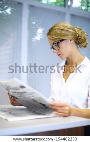 A serious girl with glasses reading a newspaper at the table in the library