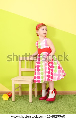 The girl in a bright dress and red shoes large size are holding the back of a chair