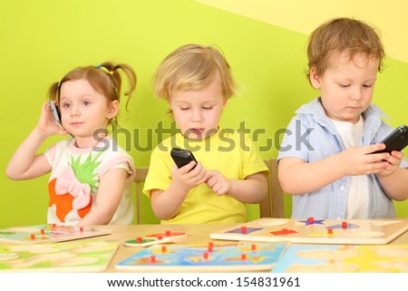 Two boys and a girl with phones in their hands are sitting at a table with toys