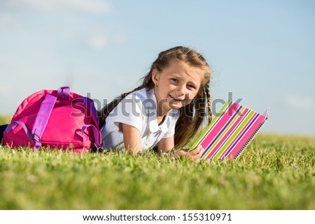 Little girl with pink backpack lying on the grass and looking in large notepad