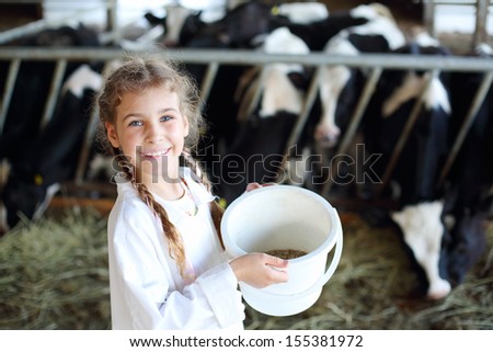 Little Beautiful Girl Holds White Bucket With Food And Smiles In Stall With Many Cows.