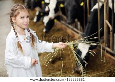 Happy cute little girl in white robe holds hay for cows in long stall.