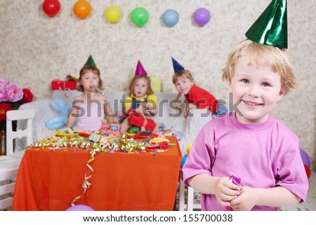 Boy in cap and with party blowers smiles at birthday party and three girls sit on couch. Inscription Happy Birthday on wall. Focus on boy.