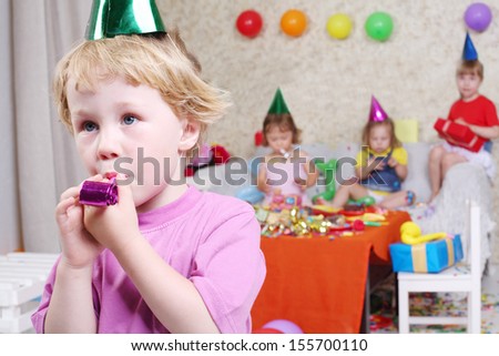 Little boy blows in party blowers at birthday party and three girls sit on couch. Inscription Happy Birthday on wall. Focus on boy.