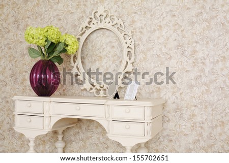 Wooden carved dressing table with blank frame and flowers in light room.