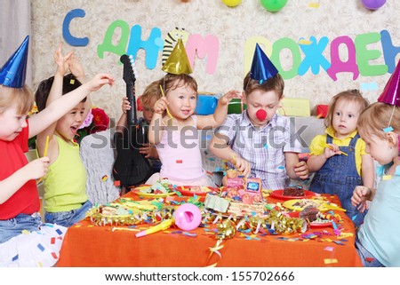 Seven happy little kids eat cake at red table at birthday party. Inscription Happy Birthday on wall.