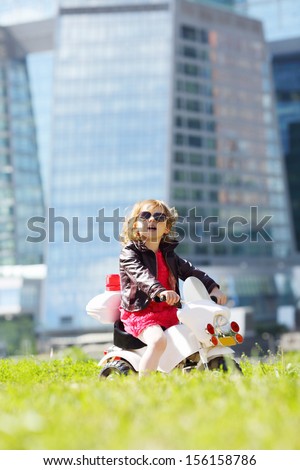 Little cute girl in leather jacket rides on toy motorbike on grass near skyscrapers at sunny day.