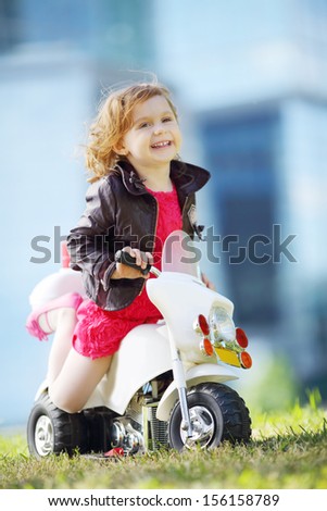 Little happy girl in leather jacket sits on toy motorbike on grass near skyscrapers.
