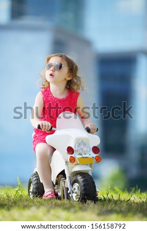 Little happy girl in sunglasses rides on toy motorbike on grass near skyscrapers.
