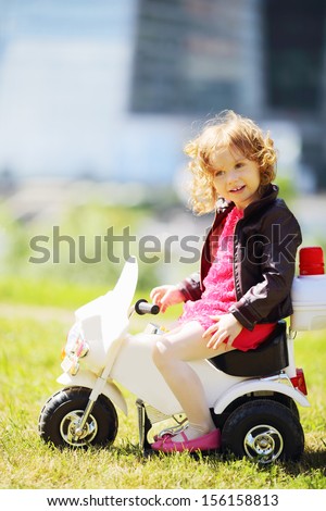 Little girl in leather jacket sits on toy motorbike on grass near skyscrapers.