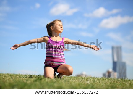 Happy little girl sits on grass on her lap, her arms outstretched and basks in sun.