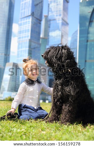 Little cute girl sits on grass with big black dog near skyscrapers at sunny day.