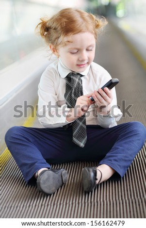 Little girl with black mobile phone sits on travelator in gallery.