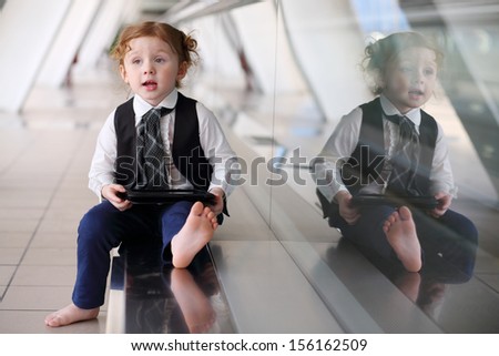 Little cute barefoot girl in tie sits on floor with tablet PC in gallery near glass wall.