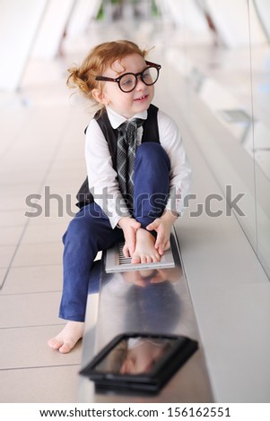 Little barefoot girl in glasses sits on floor in gallery near glass wall and looks away.