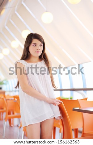 Smiling girl in white blouse and shorts stands near orange chairs in cafe.