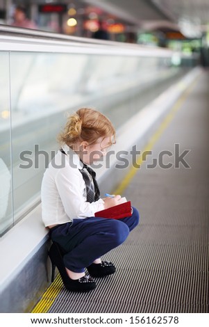 Little girl in mother shoes writes in red notebook on travelator.