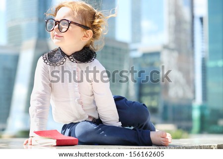 Little cute girl in glasses with red book sits on border near skys?raper at sunny day.