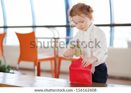 Little cute girl in white shirt stands near table in cafe and gets money from wallet.