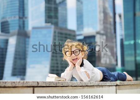 Little cute girl in glasses with book lies on border and laughs near skys?raper at sunny day.