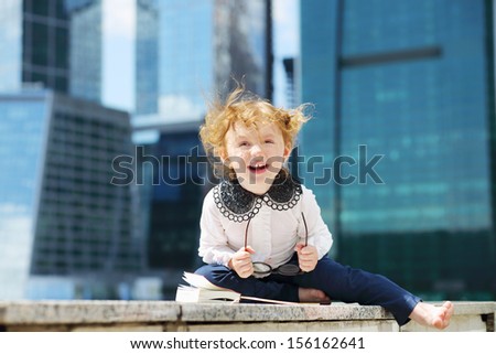 Little cute girl with book and glasses sits on border and laughs near skys?raper at sunny day.