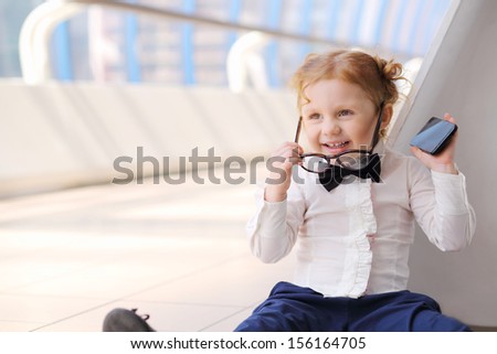 Red hair little cute girl with cell phone sits on floor and removes glasses.